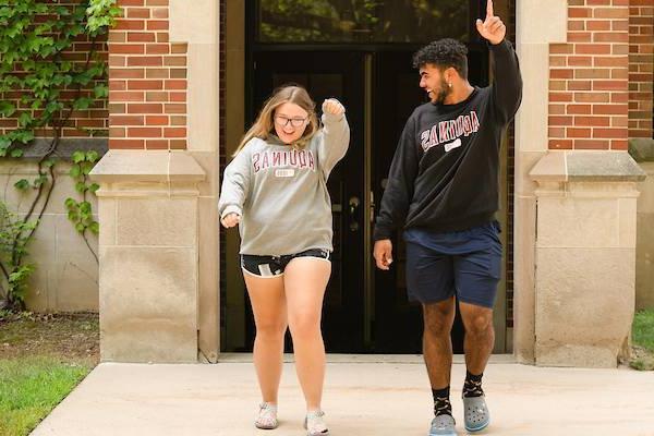 Two students in AQ sweatshirts walk with their hands in the air smiling